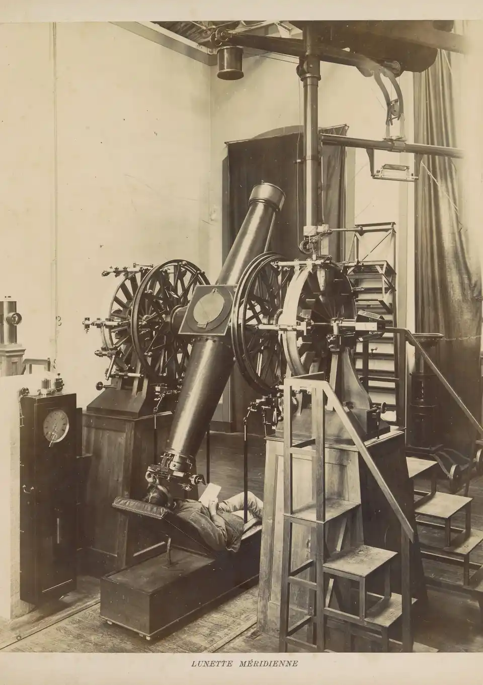 Photograph of a meridian telescope in an observatory: an observer lies on a padded couch under the eyepiece with a notebook in his hands, beside a tall pendulum clock and a wooden stepladder.